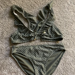 Grön prickig bikini med volangdetalj - Supersöt grön bikini med vita prickar och volangdetaljer på överdelen. Toppen har knyt framtill med små guldfärgade snäckskal som hängsmycken. Klassisk bikiniunderdel i matchande mönster. Perfekt för stranden eller poolen! Skriv om man bara vill köpa överdel/underdel separat!