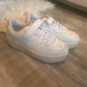 Vita sneakers med holografiska detaljer - Snygga vita sneakers med bred kardborre och snörning. Skorna har perforerad tå, platt sula och coola holografiska detaljer vid häl och kardborre. Perfekta för dig som gillar en clean look med en twist. Materialet är syntet och textil.