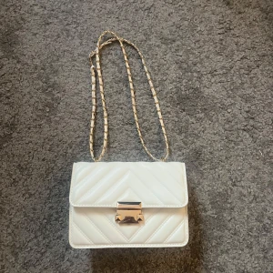 Vit quiltad axelväska med kedja - Snygg vit axelväska med quiltad design och guldfärgad kedjerem. Väskan har ett stilrent spänne framtill och är perfekt för att lyfta din outfit. Klassisk rektangulär form och trendig look. NY