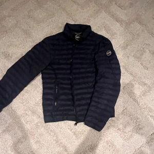 Svart dunjacka från Colmar Originals - Snygg svart dunjacka från Colmar Originals med quiltad design och två dragkedjeförsedda fickor framtill. Jackan har hög krage, diskret logga på ärmen och klassisk passform. Perfekt för kyliga dagar när du vill hålla stilen.