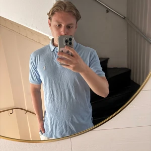 Ljusblå pikétröja från Ralph Lauren - Säljer en klassisk ljusblå pikétröja från Ralph Lauren i 100% bomull. Tröjan har korta ärmar, krage och knappar framtill samt den ikoniska broderade loggan på bröstet. Perfekt för en clean och stilren look. Sitter mer som L