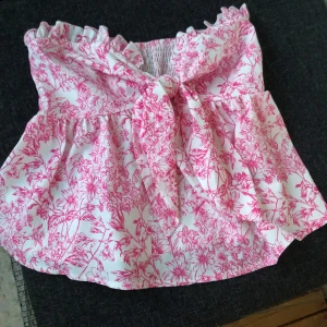 Blommig topp - Gullig vit of shoulder topp med rosa blommor på. ribbad i bak, Gummi band över brösten så den inte åker ner. Man kan knyta ifram (om ni vill ha bättre bild så tveka inte att skriva🫶🏼, de går oxå att ta bort bandet man knyter om man inte vill ha den. Tyget kan kännas lite plastigt så ni vet innan.