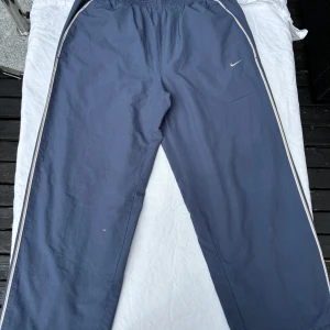 Blå Nike mjukisbyxor med vita ränder - Snygga blå mjukisbyxor från Nike med vita och gula ränder längs sidorna. Byxorna har elastisk midja med snörning och klassisk Nike-logga broderad på låret. Perfekta för chill eller träning, tillverkade i mjukt bomullsmaterial.