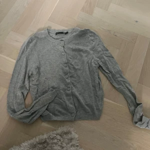 Grå kofta med knappar - En enkel och stilren grå kofta från lager 157 i storlek S. Koftan har rund halsringning, långa ärmar och knäppning framtill med matchande grå knappar. Perfekt att slänga över en t-shirt eller linne för en chill look.