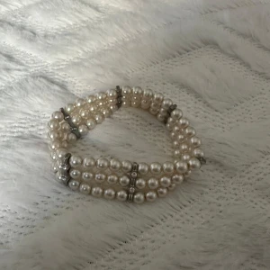Pärlarmband med strassdetaljer - Superfint armband med tre rader vita pärlor och silvriga detaljer med små glittrande stenar. Armbandet är elastiskt och sitter snyggt runt handleden. Perfekt accessoar för att lyfta din outfit med en klassisk och elegant touch.