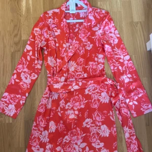Röd blommig omlottklänning gina tricot strl 38 - Snygg röd omlottklänning från Gina tricot med rosa blommigt mönster. Klänningen har långa ärmar, krage och knytband i midjan för en smickrande passform. Perfekt för dig som vill sticka ut med färg och mönster.