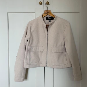 Beige ulljacka från Vero Moda - Snygg beige jacka från Vero Moda i ullblandning. Jackan har raka linjer, två stora fickor framtill och stängs med dragkedja. Enkel och clean design med rundad krage och lång ärm, perfekt för dig som gillar stilren look.