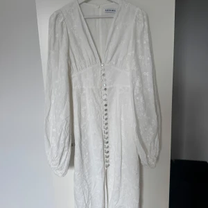 Vit långklänning från Adoore - Vit långklänning från Adoore med broderade blommor, v-ringning och långa puffärmar. Klänningen har dekorativa knappar framtill och är tillverkad i ett lätt, broderat tyg som ger en lyxig känsla. Nyskick! 