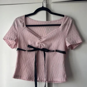 Ljusrosa T-shirt med svart rosett - Supersöt ljusrosa t-shirt med kort ärm och v-ringning från SHEIN. Toppen har en svart rosett som knyts i midjan och ett lätt skrynklat tyg som ger en cool struktur. Perfekt för sommar och festliga tillfällen! Hör gärna av dig om du vill se fler bilder eller har frågor!