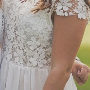 Vit blommig spetsklänning med dragkedja - Superfin vit kortklänning med blommig spets upptill och lätt, flowy kjol. Klänningen har korta ärmar och dragkedja i ryggen. Perfekt för sommarens alla tillfällen och ger en romantisk vibe.