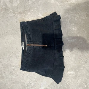 Svart jeanskjol med volang från One Teaspoon - Cool svart jeanskjol från One Teaspoon med volangdetalj upptill och synlig dragkedja framtill. Kjolen är kort och har en klassisk jeanslook med en liten läderpatch bak. Perfekt för dig som gillar edgy och trendiga plagg.