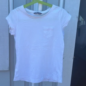 Vit t-shirt från Lindex Kids - En basic vit t-shirt från Lindex Kids i mjuk bomull. Klassisk rund halsringning, korta ärmar och en liten bröstficka framtill. Perfekt till jeans eller shorts för en clean och enkel look.