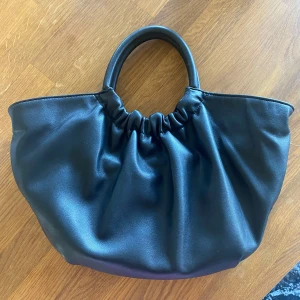 Svart handväska från MNG i skinnimitation - Snygg svart handväska från MNG i skinnimitation med rynkad design upptill och rundade handtag. Väskan har ett ljust innerfoder, innerfack med dragkedja och guldfärgade detaljer. Går att bäras i handen eller över axeln med det medföljande bandet