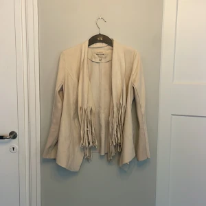 Beige mockaimitationsjacka med fransar - Säljer en beige jacka från River Island i mjuk mockaimitation med långa fransar framtill. Jackan har lång ärm, dragkedjor vid ärmslut och en snygg, lite figursydd passform. Perfekt för dig som vill ha en cool och trendig look.