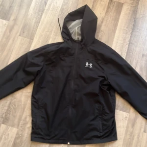 Svart vindjacka från Under Armour - Svart vindjacka från Under Armour med huva och dragkedja framtill. Jackan har en sportig look, meshfoder på insidan av huvan och logga på bröstet. Perfekt för blåsiga dagar och enkel att matcha med det mesta.