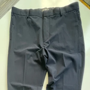 Svarta kostymbyxor H&M Slim Fit 33 - Svarta kostymbyxor från H&M i slim fit-modell. Klassisk design med pressveck, bälteshällor och två bakfickor med knapp. Snygg och stilren look som passar till skjorta eller t-shirt. Materialet är mjukt och följsamt.