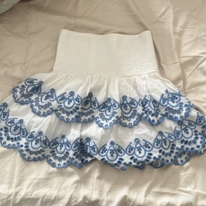 Vit kjol med blå broderi från Lindex - Supersöt vit kortkjol från Lindex med bred smock i midjan och blå broderade detaljer i vågiga lager. Kjolen har en luftig känsla och passar perfekt till varma dagar. Broderiet ger en unik och fräsch look. Kjolen har aldrig använts och är därför i super bra skick.
