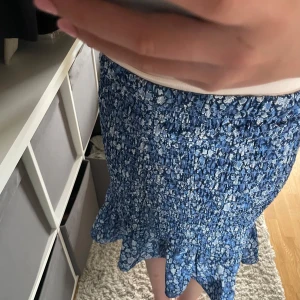 Blå blommig volangkjol - Supersöt blå kortkjol med små vita blommor och volangkant nertill. Kjolen är smockad och sitter tajt upptill men har en fin volang som ger extra rörelse. Perfekt för sommarens alla dagar och lätt att matcha med en enkel topp. Den går också att använda som en top.