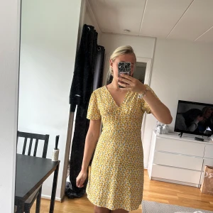 Gul blommig kortklänning med v-ringning - Superfin gul kortklänning med blommigt mönster och v-ringad hals. Perfekt för vardag.