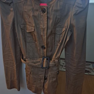 Beige kavajjacka med bälte från Promod - Snygg beige kavajjacka från Promod med markerade axlar, fyra fickor framtill och bälte i midjan. Jackan har stora knappar och axelklaffar som ger en cool look. Perfekt för dig som gillar stilrena detaljer och vill ha en jacka som sticker ut.