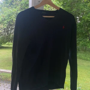 Svart v-neck tröja Polo Ralph Lauren - Svart v-ringad tröja från Polo Ralph Lauren i mjuk pima bomull. Klassisk design med röd broderad logga på bröstet och långa ärmar. Perfekt för dig som gillar stilrena och tidlösa plagg med premiumkänsla. Använd i 8 månader.