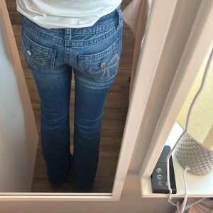Blå bootcut jeans från Gina Tricot - Säljer ett par blå bootcut jeans från Gina Tricot med låg midja. Jeansen har snygga detaljer på bakfickorna med broderade mönster och silvriga knappar. Klassisk femficksmodell i mjukt denimtyg som sitter skönt på kroppen.