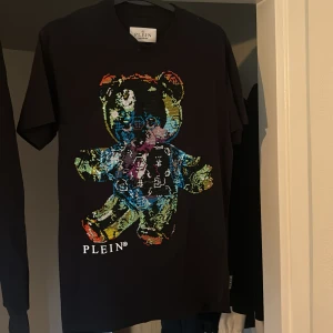Svart t-shirt med björntryck från Plein - Cool svart t-shirt från Plein med ett stort färgglatt björntryck framtill i glittriga detaljer. Klassisk passform och rund halsringning. Perfekt för dig som vill sticka ut med en unik och trendig look.