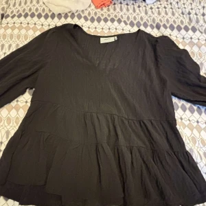 Svart volangblus från Moves - Supersnygg svart blus från Moves med v-ringning och långa ärmar. Blusen har flera volanglager som ger en flowy och trendig look. Perfekt att styla med jeans eller kjol för en chill men ändå uppklädd vibe.