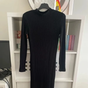 Svart ribbad långklänning - Säljer en svart långklänning med ribbad struktur och lång ärm. Klänningen har en tight passform och rund halsringning, perfekt för dig som gillar en stilren och enkel look. Materialet är stickat och känns mjukt mot huden.