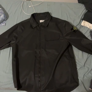 Svart overshirt från Stone Island - Säljer en svart overshirt från Stone Island med klassisk logga på ärmen. Jackan har dragkedja framtill, bröstficka med lock och lång ärm. Snygg och stilren modell som passar perfekt till streetwear eller lager-på-lager.