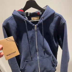Blå hoodie från Burberry med dragkedja - Snygg marinblå hoodie från Burberry med hel dragkedja och klassiskt rutigt foder i huvan. Tröjan har snörning i huvan, två fickor fram och är tillverkad i mjuk bomull. Perfekt för dig som vill ha en stilren och lyxig look med en touch av Burberrys ikoniska mönster.