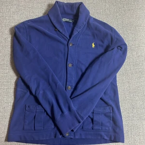 Blå kofta - Polo Ralph Lauren - Marinblå Polo Ralph Lauren kofta med svarta knappar och gult märke, koftan är i storlek L men passar också M. Vid frågor är de bara att kontakta, pris är inte hugget i sten. 