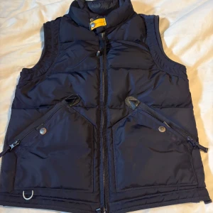 Mörkblå dunväst från Parajumpers - Snygg mörkblå dunväst från Parajumpers med hög krage, gul detalj vid halsen och två stora fickor med dragkedja och knapp. Västen har broderad logga på ryggen och coola orangea detaljer på insidan. Perfekt för lager-på-lager och streetstyle.