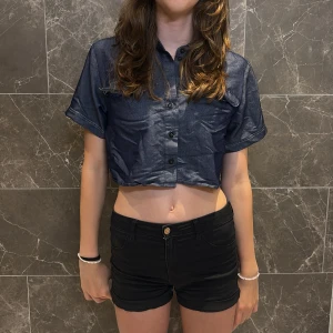 Croppad jeansskjorta från DUMA - Snygg croppad jeansskjorta från DUMA Official i mörkblå denim. Skjortan har korta ärmar, klassisk krage, knappar framtill och två bröstfickor. Perfekt för dig som gillar en trendig och avslappnad stil. Fickor där framme är lite ripped men de var så där när jag fick paketet. 