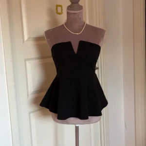 Svart peplum tubtopp - Snygg svart axelbandslös tubtopp med peplumskärning och markerad v-ringning framtill. Toppen har en figurnära passform upptill och ett utsvängt parti nedtill