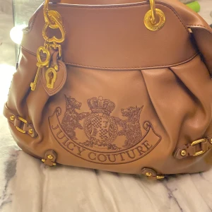 Juicy Couture brun handväska i skinn - Snygg brun handväska från Juicy Couture i skinn med broderad logga och guldfärgade detaljer. Väskan har dekorativa spännen, dragkedja och nyckelberlocker. Insidan är fodrad med Juicy Couture-tryck och en matchande liten väska ingår.