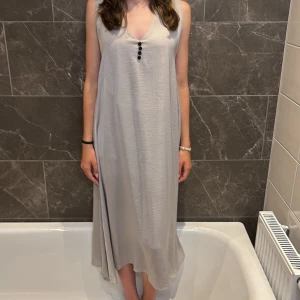 Ljusgrå ärmlös långklänning - Enkel och flowy ljusgrå långklänning med v-ringning och tre svarta knappar framtill. Klänningen är ärmlös och har en lös passform som faller snyggt. Perfekt för varma dagar när du vill ha något bekvämt men ändå stilrent.