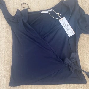 Mörkblå omlottopp från Gina Tricot - Snygg mörkblå omlottopp från Gina Tricot med långa ärmar och knyt i sidan. Toppens wrap-modell ger en schysst v-ringning och den är gjord i mjuk bomull. Perfekt att styla med jeans eller kjol för en trendig look.