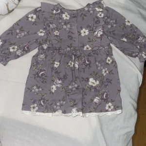 Lila blommig bebis klänning med volang - Supersöt lila kortklänning med blommigt mönster och volangdetaljer framtill. Klänningen har långa ärmar, knappar framtill och spetskant nertill. Perfekt för dig som gillar romantisk och lekfull stil. Oanvända 