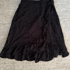 Kjol i storlek 146/152 - Fin svart långkjol i omlottmodell med knytband i midjan och volangdetaljer längs nederkanten och fram. Kjolen är i storlek 146/152 och är i ett mycket bra skick.