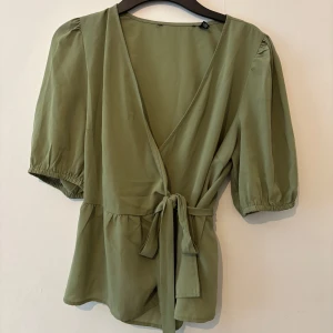 Grön sommar topp - Snygg olivgrön blus med omlottmodell och knyt i midjan. Toppen har korta puffärmar med resår och en v-ringad halsringning. Perfekt för dig som gillar en avslappnad men trendig stil.
