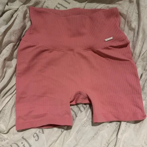 Rosa ribbade shorts från Aim'n - Supersnygga rosa ribbade shorts från Aim'n med hög midja som går att vika ner snyggt och med tight passform. Perfekta för träning eller chill dagar. Materialet är stretchigt och följsamt, tillverkat av polyamid och elastan. Diskret Aim'n-logga i metall framtill. Sitter riktigt skönt och ger en sportig vibe. Använt 1 gång 