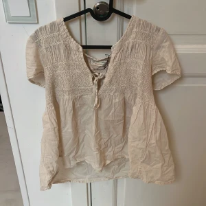 Odd Molly blus med knyt och broderi - Supersöt ljusbeige blus från Odd Molly med broderade detaljer och smock upptill. Blusen har korta ärmar, v-ringning med knyt och en luftig, flowy passform. Perfekt för dig som gillar boho-vibbar och romantisk stil.