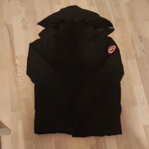 Svart dunjacka från Canada Goose - Svart dunjacka från Canada Goose med klassisk patch på ärmen och huva. Jackan är perfekt för kalla dagar och har en stilren look med diskreta detaljer. Fodrad för extra värme och har dragkedja framtill. Passar dig som vill ha både funktion och stil.