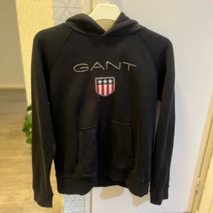 Svart hoodie från GANT med logga - Svart hoodie från GANT med broderad logga och sköld på bröstet. Klassisk känguruficka framtill, huva och långa ärmar. Mjuk bomullskänsla och sportig look, perfekt för chill dagar. Snygga detaljer med röd, vit och blå rand på insidan av kragen.