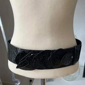 Brett svart bälte i skinn med bladdekor - Unikt svart bälte i glansigt skinn med croco-mönster och coola bladformade detaljer framtill. Bältet är brett och ger en edgy vibe till din outfit. Perfekt för att lyfta en enkel klänning eller oversized skjorta. Snyggt och statement utan att bli för mycket.