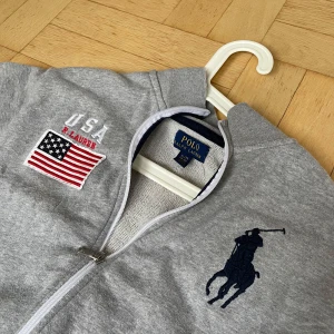 Grå ziptröja Polo Ralph Lauren USA - Grå ziptröja från Polo Ralph Lauren med broderad USA-flagga och stor marinblå polospelare på bröstet. Långärmad modell med ribbad krage och mörkblå detaljer längs axlarna. Perfekt för en sportig och avslappnad stil.