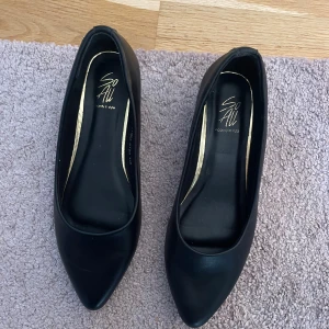 Svarta pumps med låg klack  - Stilrena svarta pumps från dinsko med spetsig tå och låg klack. Skorna är gjorda i syntetmaterial och har en klassisk, minimalistisk design med en liten guldfärgad detalj bak. Perfekta för dig som gillar en clean look med lite edge. Använda 1 gång 
