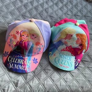 Frozen kepsar med Elsa och Anna - Två färgglada kepsar med motiv av Elsa och Anna från Frozen. Ena kepsen har texten 'Celebrate Summer' i rosa, lila och blå toner, den andra 'Sister Queens' i turkos, rosa och lila. Båda har blommiga detaljer och justerbar baksida. Perfekt för dig som älskar Disney!