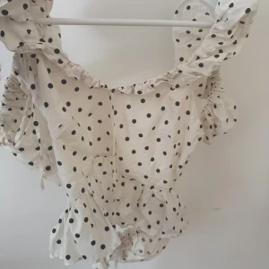 Prickig volangblus från H&M - Supersöt blus från H&M i vitt med svarta prickar. Toppen har volangdetaljer vid axlar och ärmslut, samt en lätt rynkad midja och korta puffärmar. Perfekt för dig som gillar en lekfull och trendig stil.